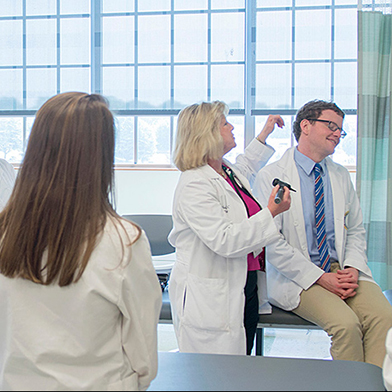 A blonde woman in white coat looks into the ear of a man wearing a white coat who is sitting on an exam table.